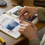 Person creating a travel scrapbook with photos, washi tape, and handwritten notes in a journal